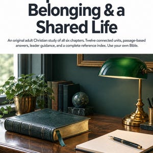 May include: A book titled Ephesians: Belonging & a Shared Life with a dark green leather cover and gold-edged pages. The book sits on a wooden desk next to a green banker lamp, a fountain pen, a notebook, and a potted plant.