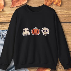 Op de afbeelding: Een zwart sweatshirt met lange mouwen en een grafische print van een wit spook, een oranje pompoen met een gestikte mond en een beige schedel in een horizontale rij op de borst, voorzien van geribbelde boorden en een ronde halslijn.