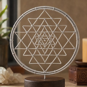 May include: A silver-colored wire Sri Yantra sculpture featuring a complex geometric pattern of interlocking triangles within a circular frame, mounted on a solid, dark brown cylindrical wooden base. The metal structure is crafted from thin wire.