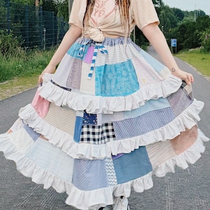 May include: A long, tiered patchwork skirt featuring various blue, white, and pink fabric patterns, including gingham, stripes, and paisley. The skirt has a drawstring waist and multiple horizontal white ruffled layers, paired with a beige t-shirt.