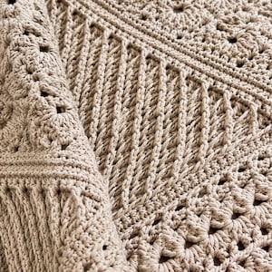 May include: A beige crochet blanket featuring a combination of intricate floral-inspired square motifs and sections of vertical ribbed stitching. The textile displays a consistent light tan color throughout with a textured, hand-crafted appearance.