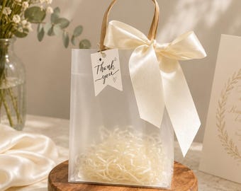 May include: A translucent frosted gift bag with a cream-colored satin ribbon bow tied to the handle. A small white rectangular tag hangs from the handle with the text "Thank you" written in black script, accompanied by a small star and heart icon.