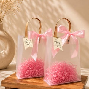 May include: Two clear frosted gift bags with brown handles, each decorated with a pink satin ribbon bow and a small white tag featuring the printed text Thank you. The bags are filled with pink shredded paper filler for gift presentation.