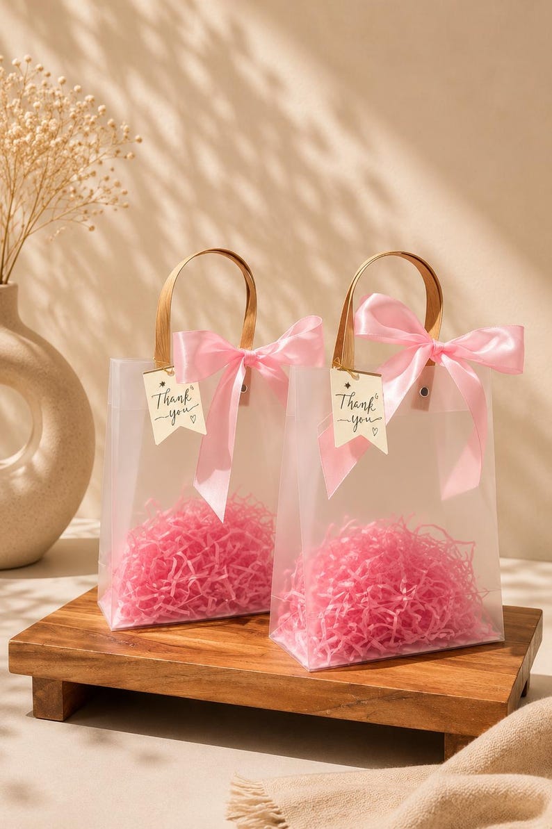 May include: Two clear frosted gift bags with brown handles, each decorated with a pink satin ribbon bow and a small white tag featuring the printed text Thank you. The bags are filled with pink shredded paper filler for gift presentation.