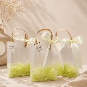 May include: Three clear, frosted gift bags with light brown handles and cream-colored ribbons tied in bows. Each bag contains light green shredded paper filler and features a small white tag with the text Thank you printed in black ink.