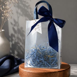 May include: A translucent gift bag with navy blue ribbon handles and a matching bow. The bag contains blue shredded paper filler and features a white tag with the text thank you and a small heart symbol attached to the handle.