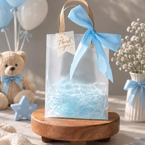 May include: A translucent frosted gift bag with light blue paper shred filler, featuring a light blue satin ribbon bow on the handle and a small white tag with the text Thank you written in brown script, centered on a round wooden pedestal.