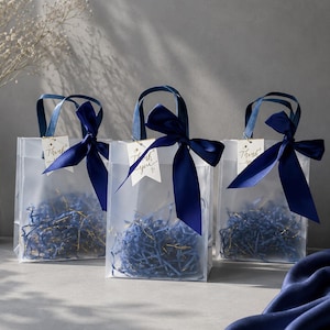 May include: Three translucent frosted gift bags with dark blue satin ribbon bows tied to the handles. Each bag contains blue shredded paper filler and features a small white tag with the handwritten text Thank you and a small heart icon.