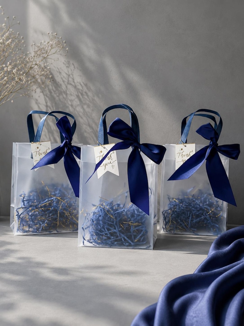 May include: Three translucent frosted gift bags with dark blue satin ribbon bows tied to the handles. Each bag contains blue shredded paper filler and features a small white tag with the handwritten text Thank you and a small heart icon.