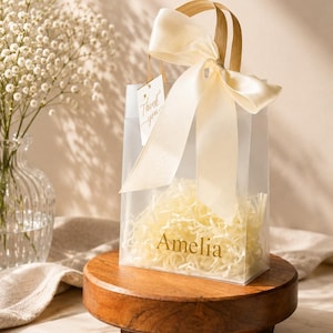May include: A clear, rectangular gift bag featuring the name Amelia printed in gold lettering on the front. The bag is filled with light-colored shredded paper filler and is tied with a large, cream-colored satin ribbon bow. A small tag attached to the handle reads Thank you.