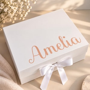 May include: A square white gift box featuring the name Amelia printed in a metallic rose gold script font on the lid. The box is secured with a white satin ribbon tied in a bow at the front center, providing a clean and elegant presentation.