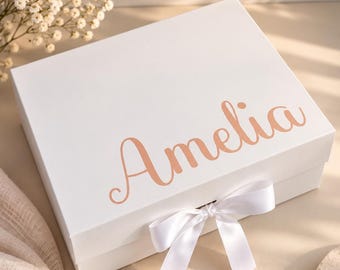 May include: A square white gift box featuring the name Amelia printed in a metallic rose gold script font on the lid. The box is secured with a white satin ribbon tied in a bow at the front center, providing a clean and elegant presentation.