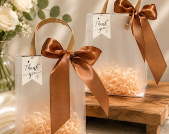 May include: Two clear rectangular gift bags with brown satin ribbon bows and tan paper filler. Each bag features a white tag with the text Thank you and a small heart symbol. The bags have sturdy handles and a transparent design for visibility.