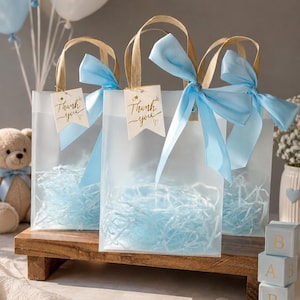 May include: Three translucent frosted gift bags with gold handles and light blue satin ribbons. Each bag contains light blue shredded paper filler and features a small white tag with the gold cursive text Thank you and a heart symbol.