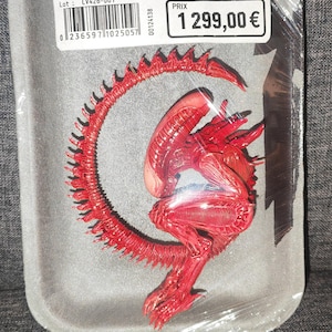Puede incluir: Una figura de Xenomorfo roja empaquetada en una bandeja de plástico gris con una película transparente. La etiqueta dice BOUCHERIE DU TERROIR, XÉNOMORPHE ADULTE PRÊT À L'EMPLOI, Origine: LV-426, Lieu de capture: LV-426, Lieu de découpe: Station Sevastopol, 102,5kg.