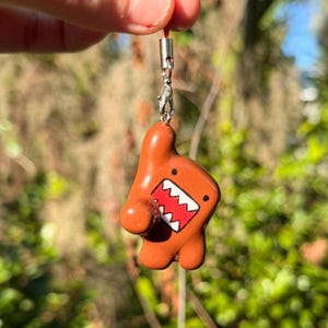 May include: A brown Domo-kun character charm featuring a wide open mouth with jagged white teeth and a red interior. The figure is attached to a silver-toned metal lobster clasp and a thin red string loop for hanging as a decorative accessory.