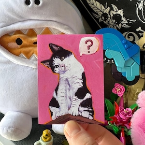 May include: A rectangular art print featuring a painting of a black and white cat sitting on a brown ledge against a solid pink background. The cat is looking forward with a tilted head, and a white speech bubble containing a red question mark is visible.