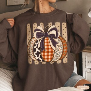 Könnte beinhalten: Ein dunkelbraunes Sweatshirt mit Rundhalsausschnitt und einem großen Kürbis-Motiv mit violetter Schleife. Der Kürbis ist in Segmente mit Kuhflecken-, orangefarbenem Vichy-, Blumen- und Karomustern unterteilt, vor einem Hintergrund aus vertikalen Streifen.