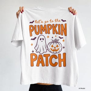 Könnte beinhalten: Ein weißes T-Shirt mit einem Grafikdruck und dem orangefarbenen Text "let's go to the PUMPKIN PATCH". Das Design zeigt ein weißes Gespenst, einen orangefarbenen Kürbis, kleine orangefarbene Sterne und dunkelviolette Fledermäuse.