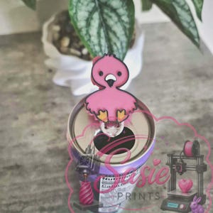 May include: A pink flamingo-shaped beverage can tab opener with a black outline, black eyes, and yellow feet. The opener is attached to the top of a metal soda can. A pink logo with the text Sasie PRINTS is overlaid on the lower portion of the image.