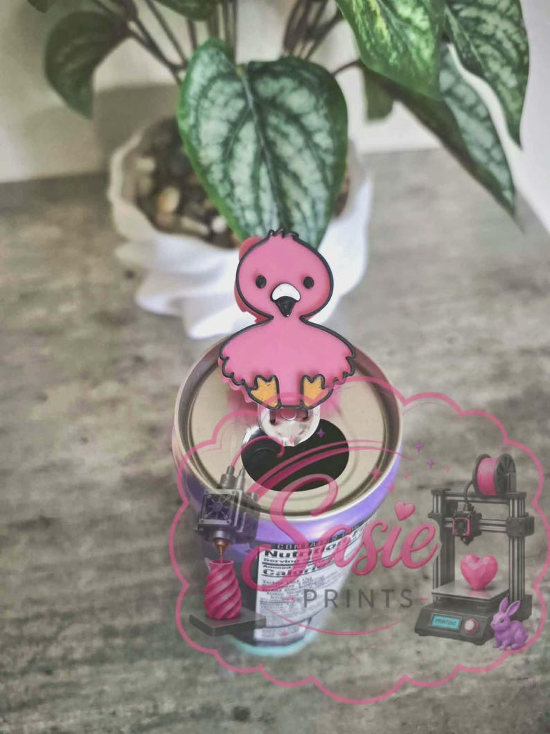 May include: A pink flamingo-shaped beverage can tab opener with a black outline, black eyes, and yellow feet. The opener is attached to the top of a metal soda can. A pink logo with the text Sasie PRINTS is overlaid on the lower portion of the image.