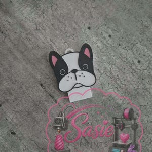 May include: A white and black French bulldog head charm with pink inner ears and a small loop at the top. Below the charm is a pink logo featuring the text Sasie Prints, accompanied by illustrations of a 3D printer, a heart, and a rabbit.