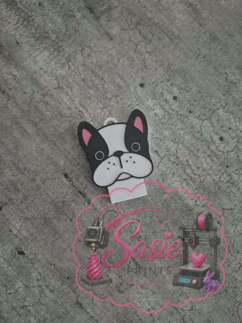 May include: A white and black French bulldog head charm with pink inner ears and a small loop at the top. Below the charm is a pink logo featuring the text Sasie Prints, accompanied by illustrations of a 3D printer, a heart, and a rabbit.