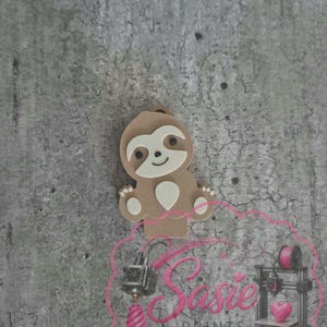 May include: A light brown sloth-shaped object with a cream-colored face and belly patch, featuring small black eyes and a smiling mouth. Below the sloth is a pink logo with the text Sasie PRINTS, featuring illustrations of 3D printers.