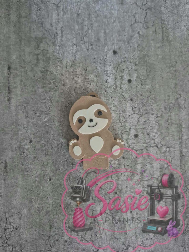 May include: A light brown sloth-shaped object with a cream-colored face and belly patch, featuring small black eyes and a smiling mouth. Below the sloth is a pink logo with the text Sasie PRINTS, featuring illustrations of 3D printers.