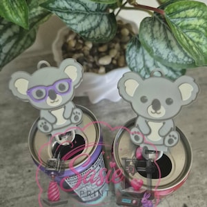 May include: Two gray koala-shaped soda can tabs, one featuring purple glasses, designed to fit onto the pull tab of a beverage can. The koalas are light gray with white accents on the ears and belly, and each has a small loop at the top.