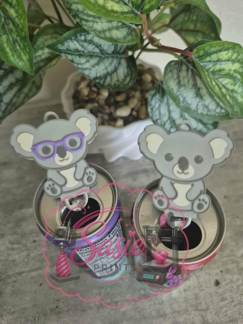 May include: Two gray koala-shaped soda can tabs, one featuring purple glasses, designed to fit onto the pull tab of a beverage can. The koalas are light gray with white accents on the ears and belly, and each has a small loop at the top.