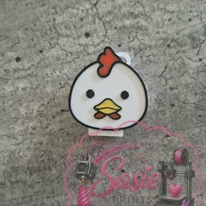 May include: A white chicken-shaped charm with a red comb, black eyes, and a yellow beak. The charm features a black outline and a small white loop at the top for attachment. A pink watermark with the text Sasie PRINTS is visible at the bottom.