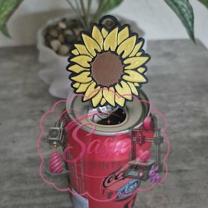 May include: A yellow and brown sunflower-shaped plastic tab opener attached to the top of a red aluminum soda can. The sunflower has a black outline and a small circular hole at the top. A pink logo with the text Sasie Prints is overlaid.