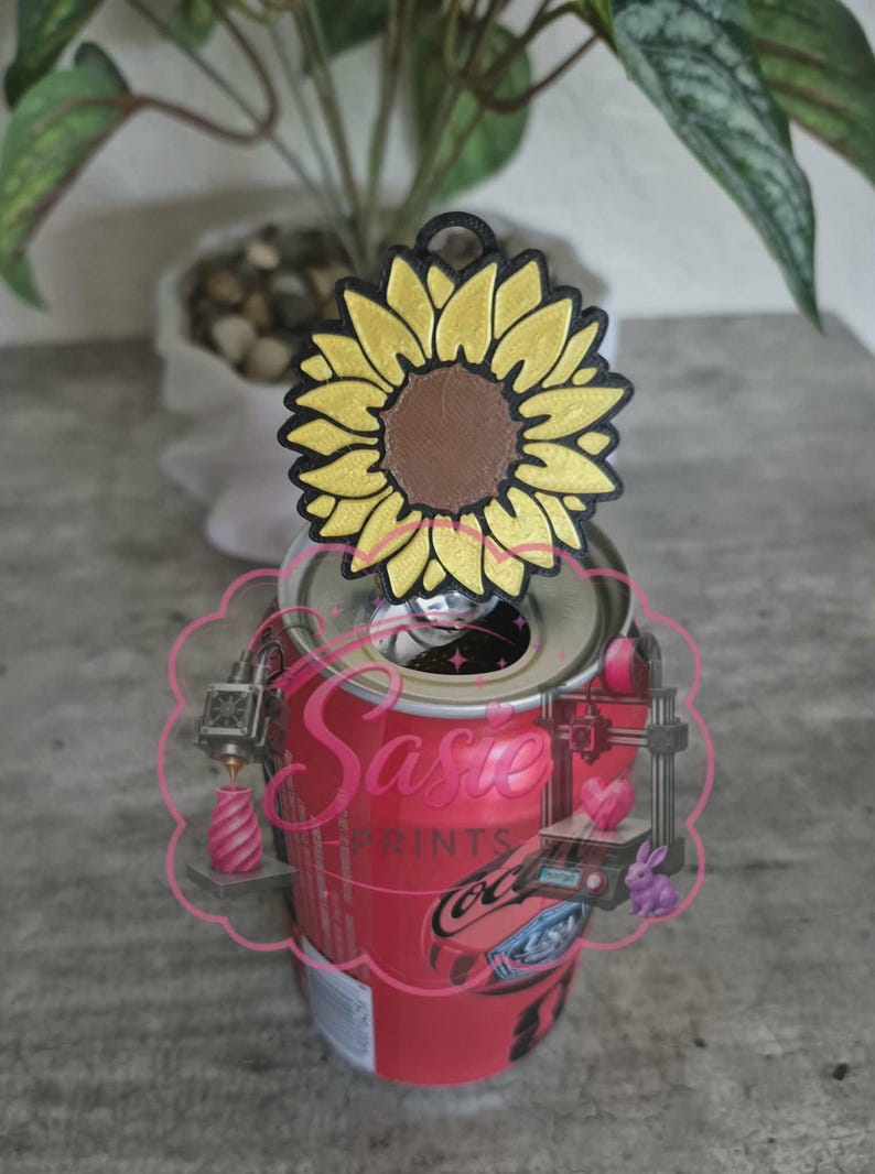 May include: A yellow and brown sunflower-shaped plastic tab opener attached to the top of a red aluminum soda can. The sunflower has a black outline and a small circular hole at the top. A pink logo with the text Sasie Prints is overlaid.