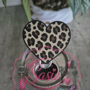 May include: A heart-shaped topper with a tan and black leopard print pattern. The topper is attached to a circular metallic soda can tab. A pink graphic overlay features the text "Sasie" in a cursive font, along with 3D printer illustrations.