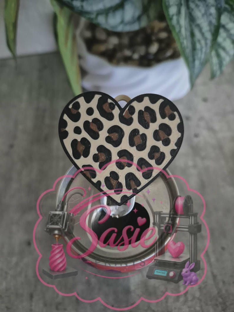 May include: A heart-shaped topper with a tan and black leopard print pattern. The topper is attached to a circular metallic soda can tab. A pink graphic overlay features the text "Sasie" in a cursive font, along with 3D printer illustrations.