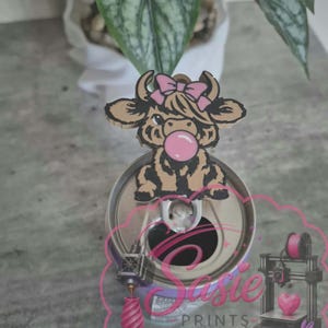 May include: A brown and black highland cow topper with a pink bow and bubblegum, attached to a silver metal soda can tab. The image features a pink logo with the text Sasie PRINTS, a 3D printer graphic, a pink heart, and a small purple rabbit.
