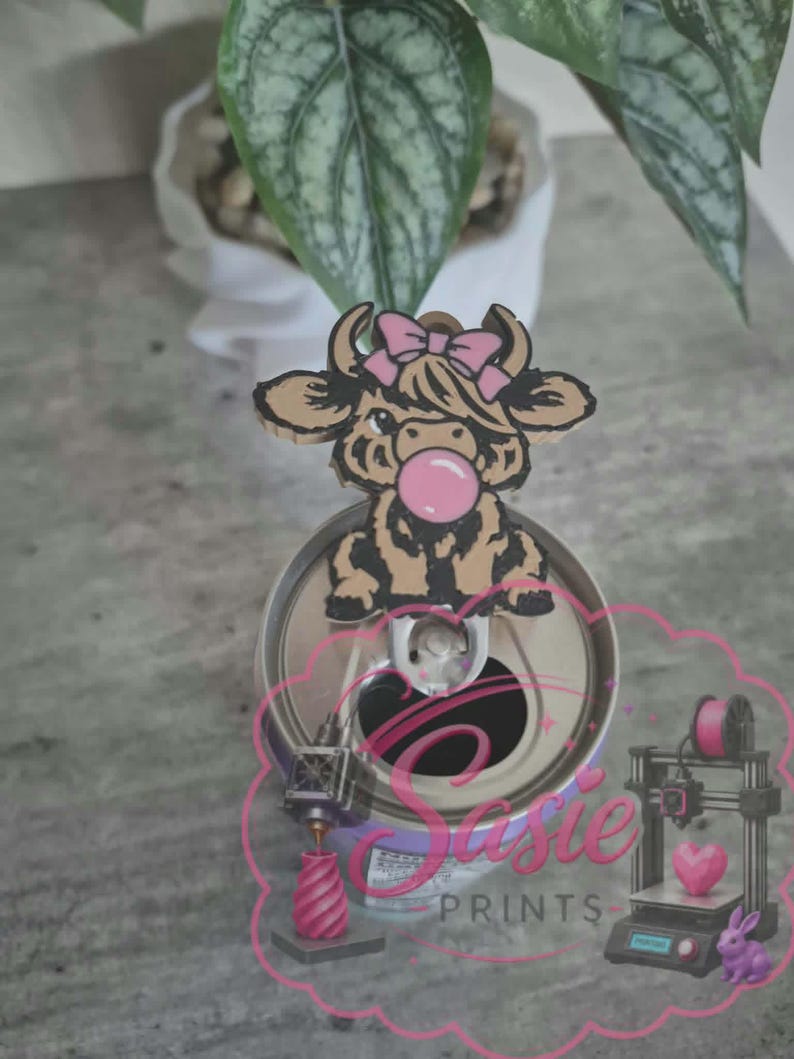 May include: A brown and black highland cow topper with a pink bow and bubblegum, attached to a silver metal soda can tab. The image features a pink logo with the text Sasie PRINTS, a 3D printer graphic, a pink heart, and a small purple rabbit.