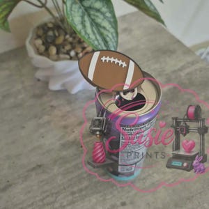 May include: A brown and white American football-shaped topper attached to the pull tab of a beverage can. The can features a nutrition facts label and a pink logo graphic with the text "Susie PRINTS" depicting a 3D printer, a heart, and a rabbit.