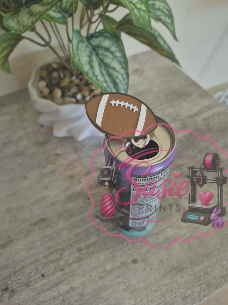 May include: A brown and white American football-shaped topper attached to the pull tab of a beverage can. The can features a nutrition facts label and a pink logo graphic with the text "Susie PRINTS" depicting a 3D printer, a heart, and a rabbit.