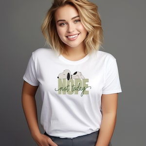 May include: A white short-sleeved t-shirt featuring a graphic of a sleeping dog character resting on top of the block-lettered text NOPE, with the cursive script text not today layered over it in a dark green color.