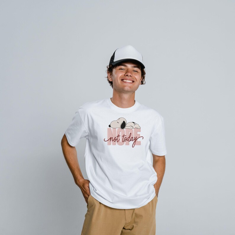 May include: A white short-sleeved t-shirt featuring a graphic of a sleeping dog above the text "nope not today" in a script font, paired with a white and black trucker hat and tan pants. The graphic includes a light pink rectangular background.