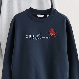 May include: A navy blue crewneck sweatshirt featuring the white text "OFF" in block letters followed by the cursive word "line" and a red stitched heart outline. The garment has long sleeves and a ribbed neckline, with a light grey fabric visible behind.