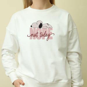 May include: A cream-colored crewneck sweatshirt featuring a graphic of a cartoon dog lying down above the word NOPE in large, light pink block letters, with the words not today written in a dark red cursive script layered over the text.