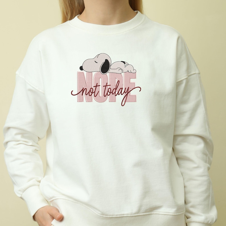 May include: A cream-colored crewneck sweatshirt featuring a graphic of a cartoon dog lying down above the word NOPE in large, light pink block letters, with the words not today written in a dark red cursive script layered over the text.