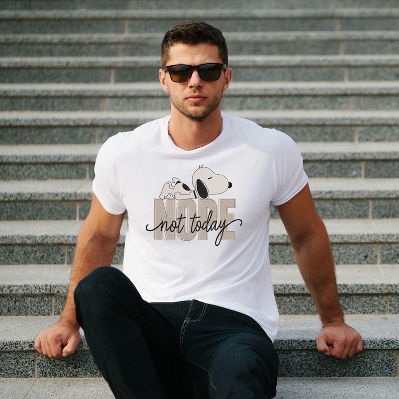 May include: A white short-sleeved t-shirt featuring a graphic of a cartoon dog lying down above the text NOPE in block letters and the cursive script text not today. The shirt has a crew neckline and is made of a plain white fabric material.