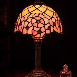 Peut inclure: Une lampe de table en vitrail avec un abat-jour en forme de dôme orné de motifs floraux roses et d'accents rouge foncé. La lampe possède un pied en métal couleur bronze foncé avec une colonne cannelée et un socle circulaire décoratif.
