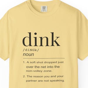 May include: A pale yellow short-sleeve t-shirt featuring black text that defines the word dink as a soft shot dropped over the net into the non-volley zone and the reason you and your partner are not speaking, with a Comfort Colors brand label.