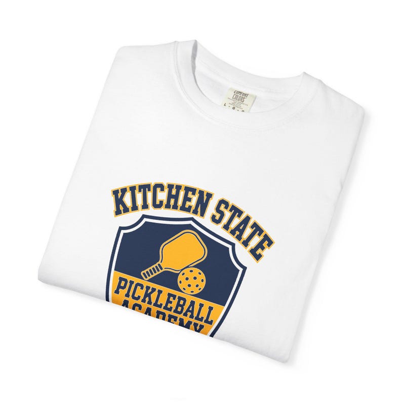 Może przedstawiać: Biały t-shirt z kr&oacute;tkim rękawem z granatowo-ż&oacute;łtą grafiką na piersi. Wz&oacute;r zawiera emblemat w kształcie tarczy z rakietką i piłeczką do pickleballa oraz napis KITCHEN STATE PICKLEBALL ACADEMY wykonany pogrubioną czcionką.