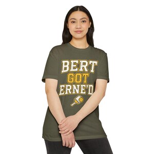 May include: An olive green short-sleeve t-shirt featuring the text "BERT GOT ERNE'D" in a distressed white and yellow font. Below the text is a graphic of a yellow and white pickleball paddle with a ball, centred on the chest of the shirt.
